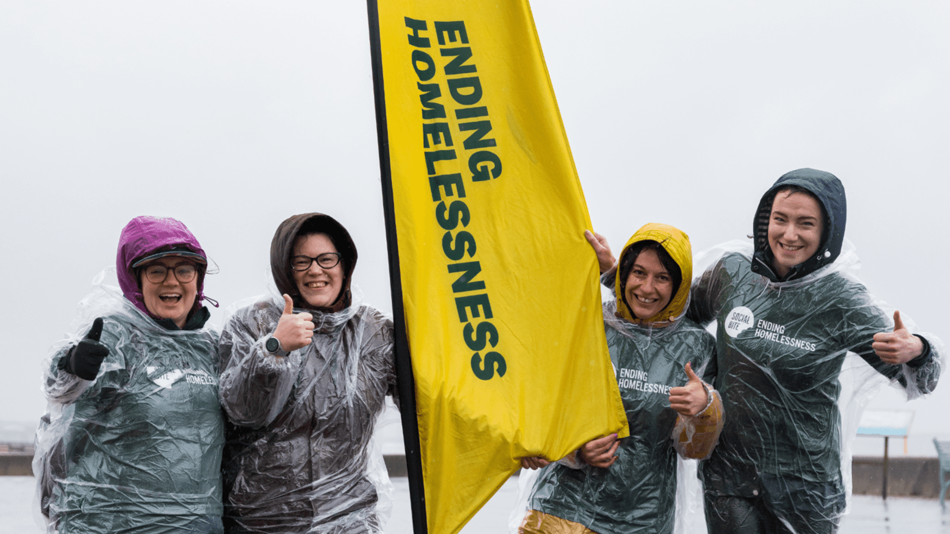 Four women in raincoats and clear ponchos smile and give thumbs up in very wet weather. They have a tall yellow banner between them that reads 'ending homelessness'