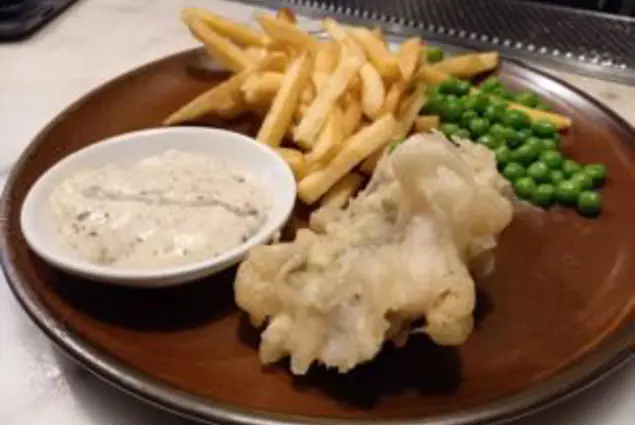 A brown plate with battered fish, chips, peas and a bowl of tartare sauce