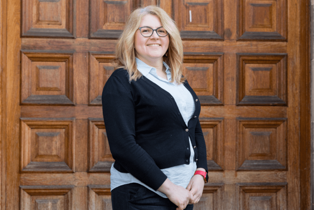 A woman with blonde hair and glasses stands smiling in front of a wooden door