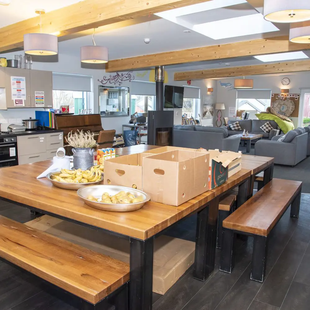 A huge communal kitchen dining table with boxes and snacks on it, looking on to a living room