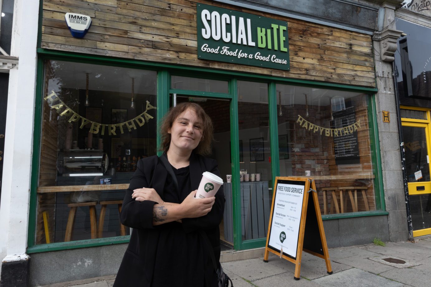A woman holding a takeaway coffee cup standing with arms crossed outside a shop called Social BIte