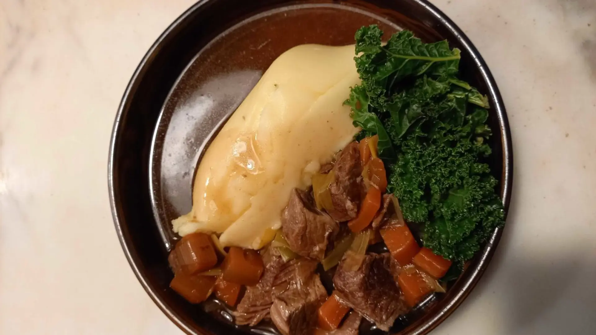 A round bowl with mashed potato, kale, beef stew and gravy