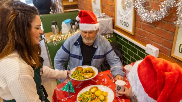 Two men in Santa hats sitting at a table with a red tablecloth receive a plate of Christmas dinner from a woman in a green apron.