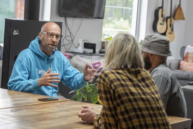 A bald man with glasses and a bright blue hoody talks to a a woman and a man across a wooden table